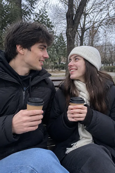 A couple in winter coats sharing a coffee on a park bench