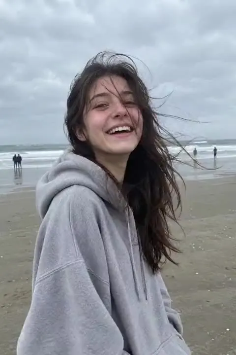 A young woman in a grey hoodie laughing on a windy beach, a still photo