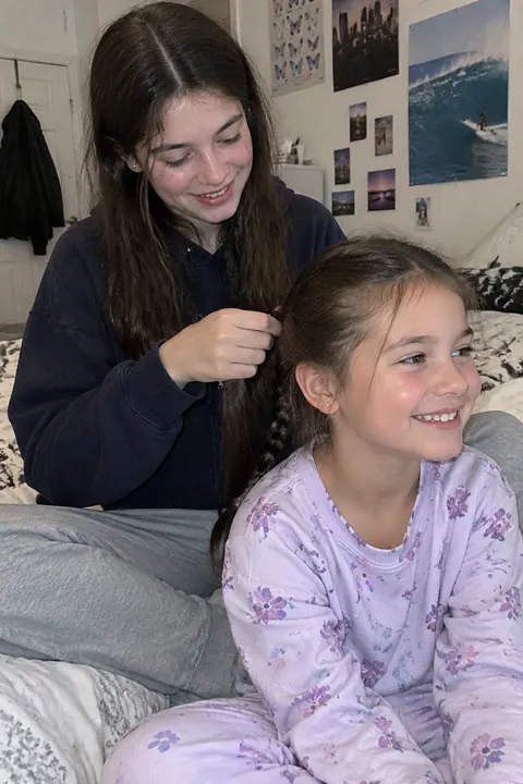 An older girl sitting on a bed braiding a younger smiling girl's hair