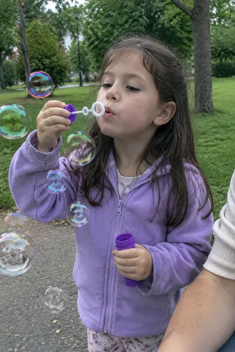 A young girl in a purple fleece jacket blowing soap bubbles outdoors