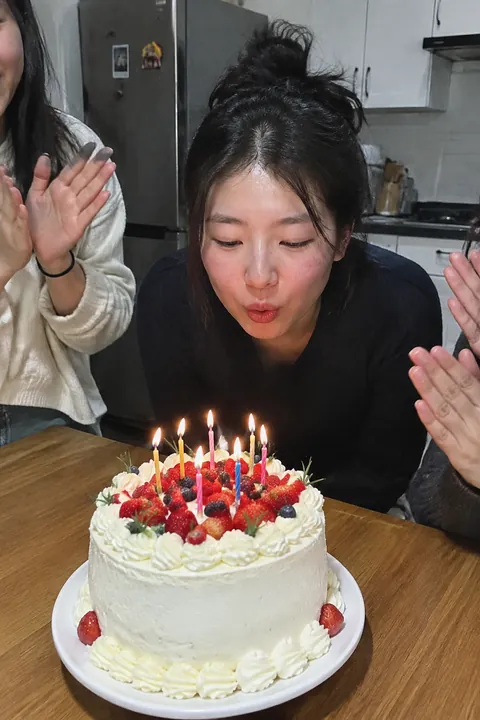 A young woman leaning over a lit birthday cake surrounded by clapping friends