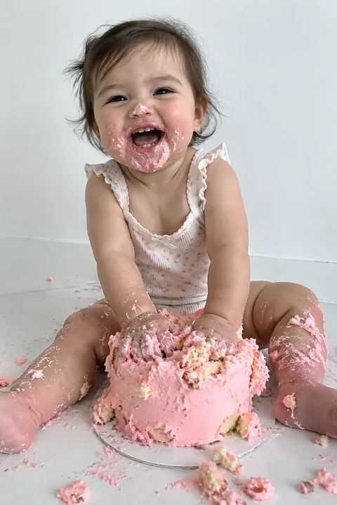 A laughing baby covered in pink frosting sitting before a smashed cake