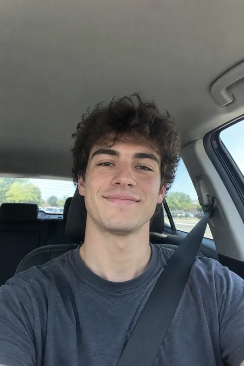A young man with curly hair taking a selfie in the driver's seat of a car