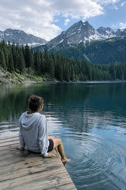 A woman sitting on a wooden dock dips her feet into a mountain lake