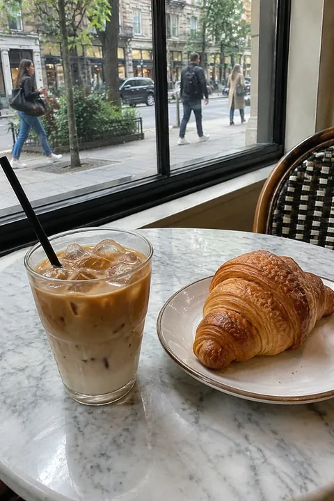 An iced coffee and a croissant on a marble table by a window