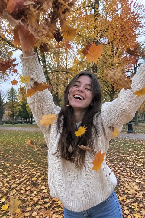 Smiling young woman in a cream sweater throwing autumn leaves in a park