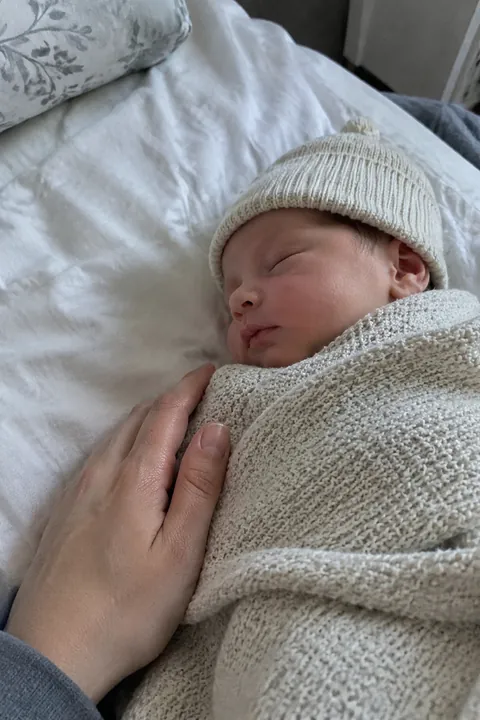 A sleeping newborn wrapped in a knit blanket next to a caregiver's hand