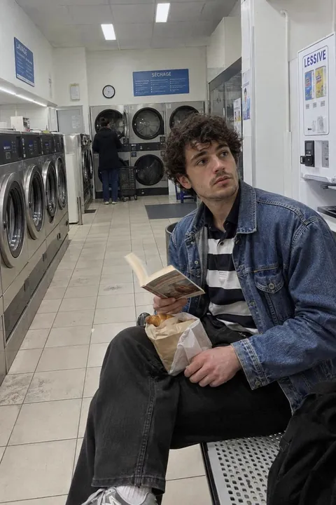 A man in a denim jacket reading a book on a bench in a laundromat