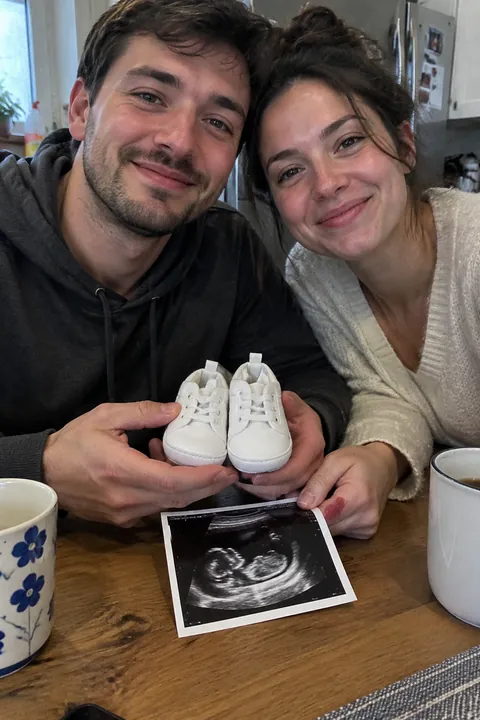 A smiling couple sitting at a table holding baby shoes and an ultrasound picture