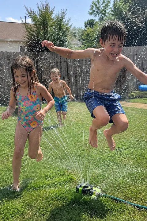 Three young children playing and jumping around a lawn sprinkler on a sunny lawn