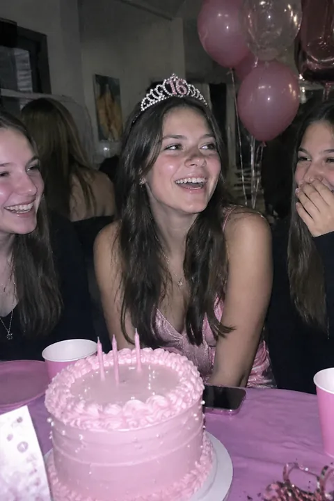 A birthday girl in a tiara sits smiling behind a pink cake with candles