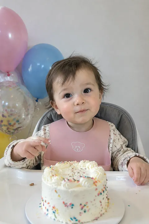 A one-year-old in a high chair with a small birthday cake and balloons