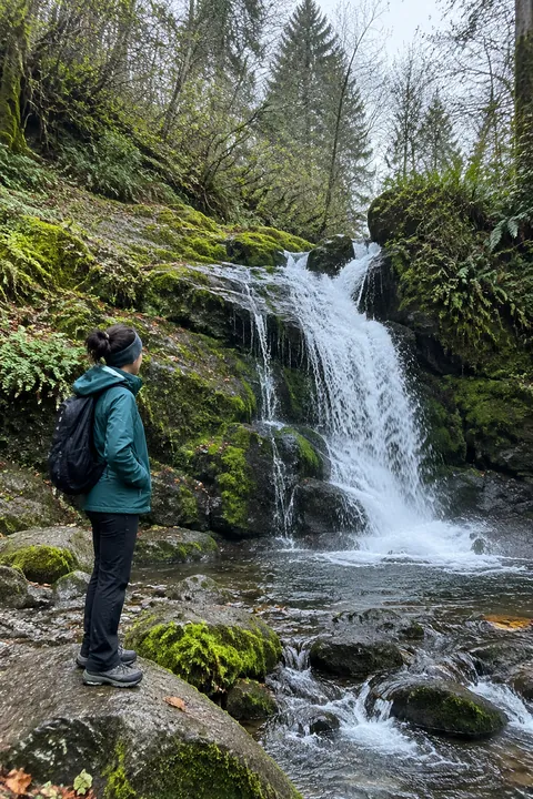 A hiker standing on mossy rocks looking at a forest waterfall