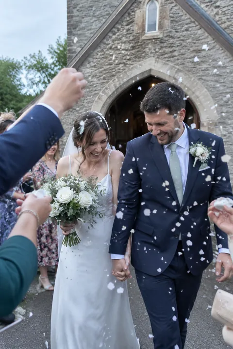 A bride and groom smile while walking through confetti outside a stone church